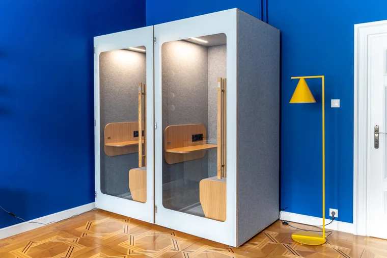 Two light HIDE H1 acoustic booths in an office with deep blue walls and a yellow lamp