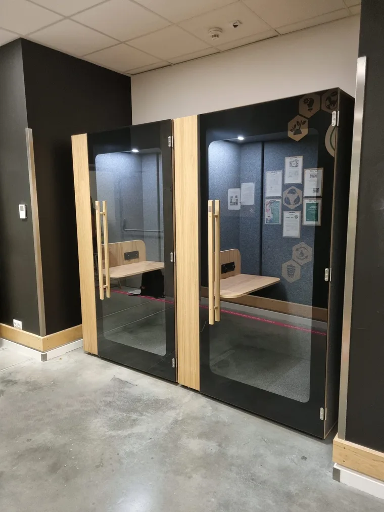 Two black HIDE H1+ acoustic booths in a staff area with eco-friendly icons and certificates