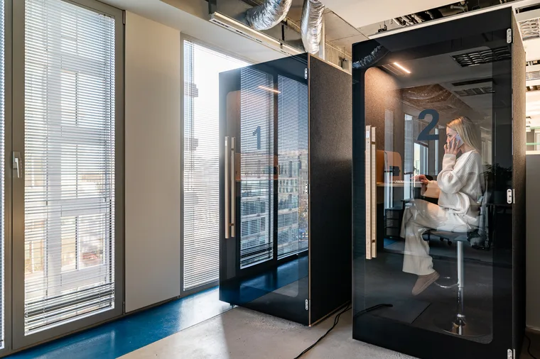 Two black HIDE H1 booths by a window; in booth 2 a woman is talking on the phone