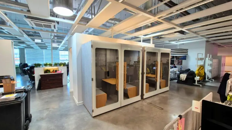 Two light HIDE H2 acoustic booths in a warehouse-style office with concrete floor and exposed ceiling ducts