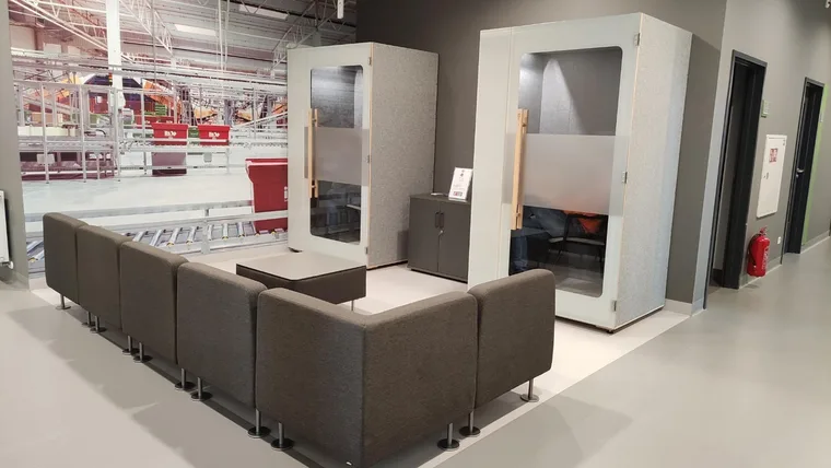 Two light-grey HIDE H1+ acoustic booths in a waiting area with a corner sofa and a warehouse mural