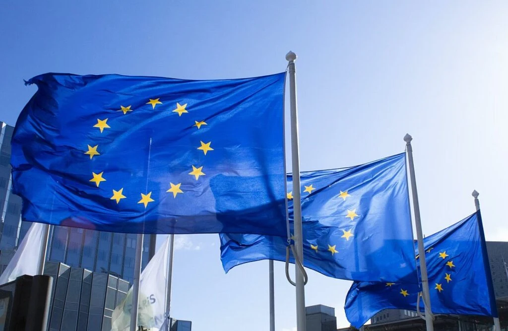 Three European Union flags on masts in front of a modern building under a blue sky