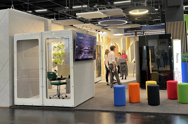 HIDE Mini Office booth at a trade fair stand, with a group of visitors and colourful stools behind